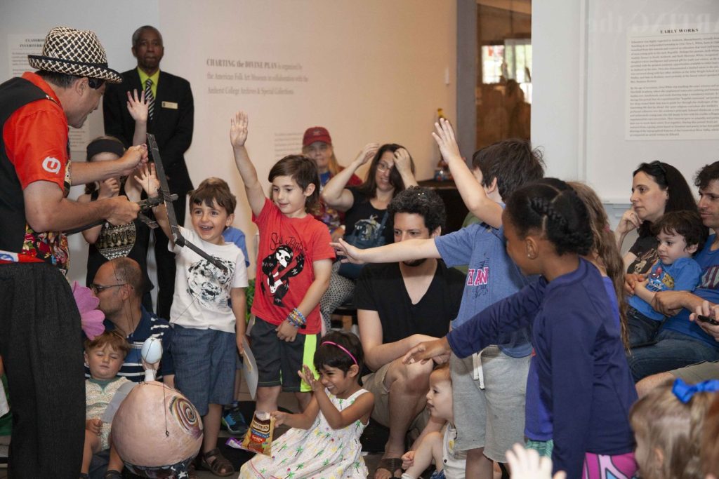 a group of excited children surround an artist and interact with his piece of art that involves wood and string