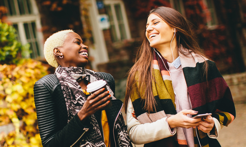 two women laugh and walk with a cup of coffee in stylish fall outfits
