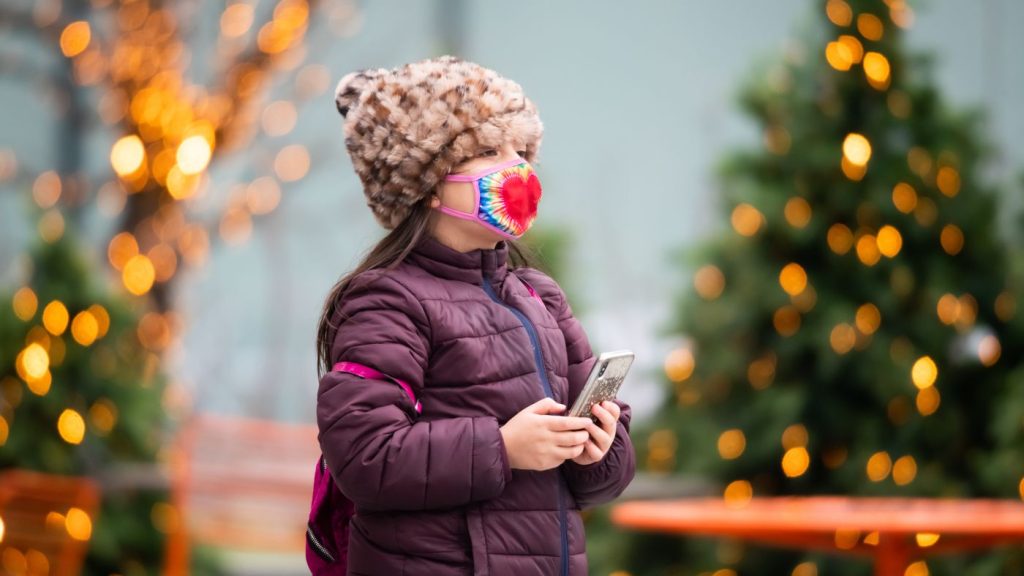 Getty Images - little girl stands in front of holiday lights outside in a winter coat and a face mask