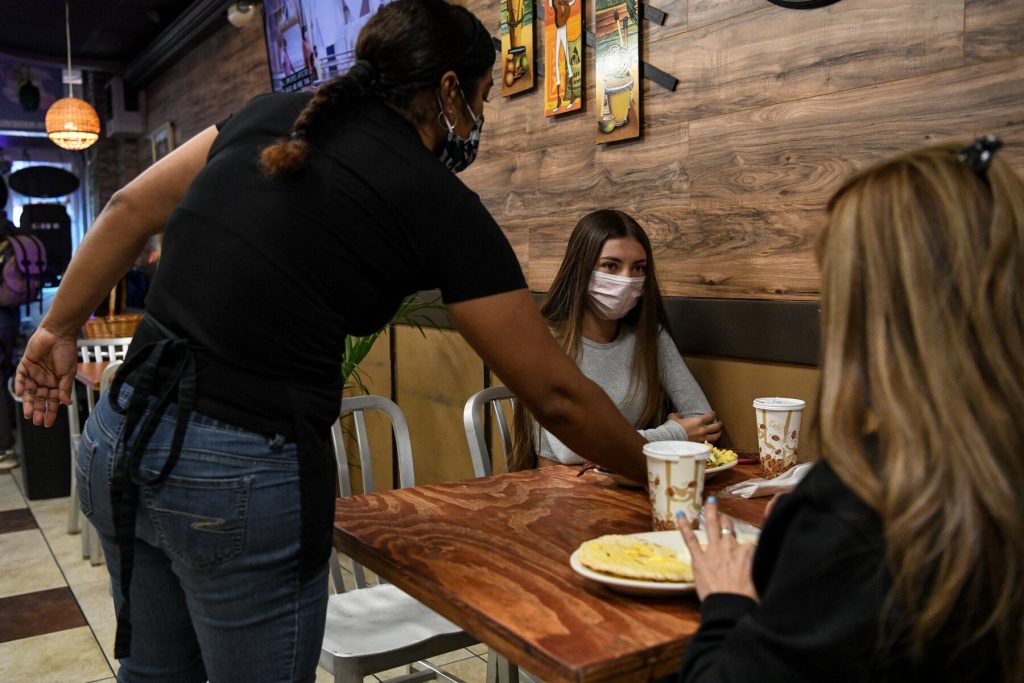 a cafe server puts down a plate of food in front of a customer seated inside wearing a mask