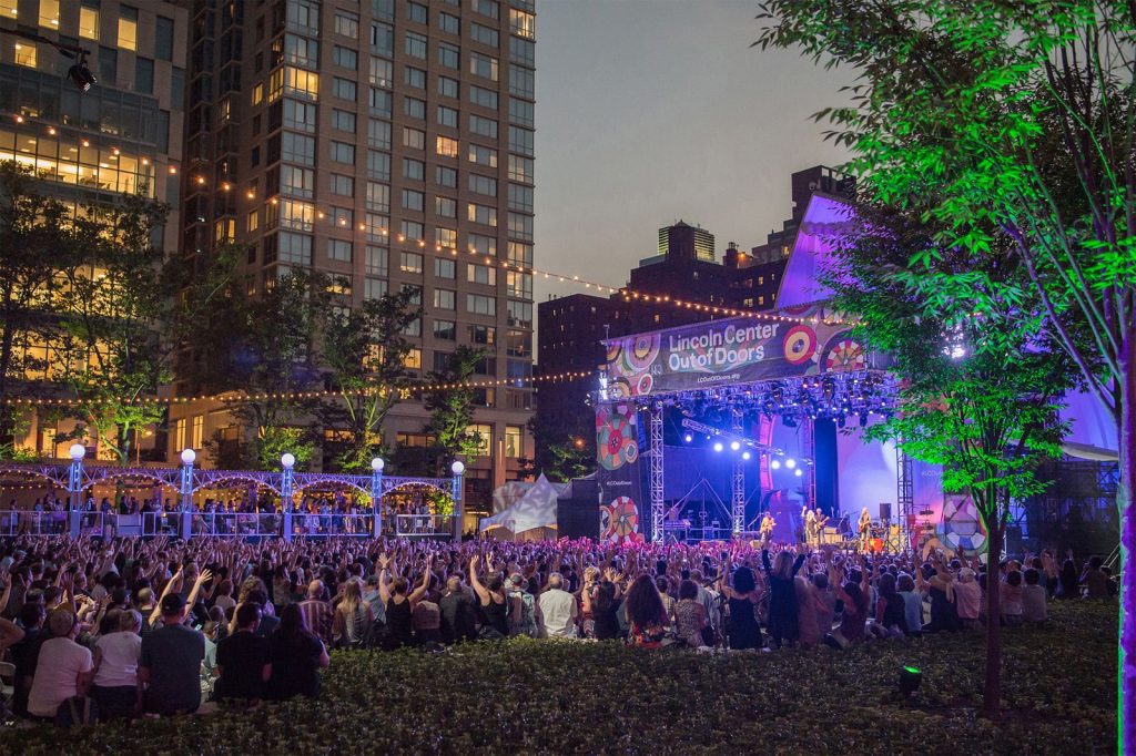 a large crowd of people enjoy a musical performance in Damrosch Park at night