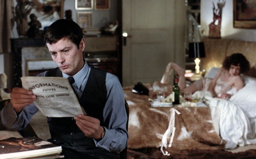 a still from Monsieur Klein--Klein reads the paper while a woman lounges in bed in the background