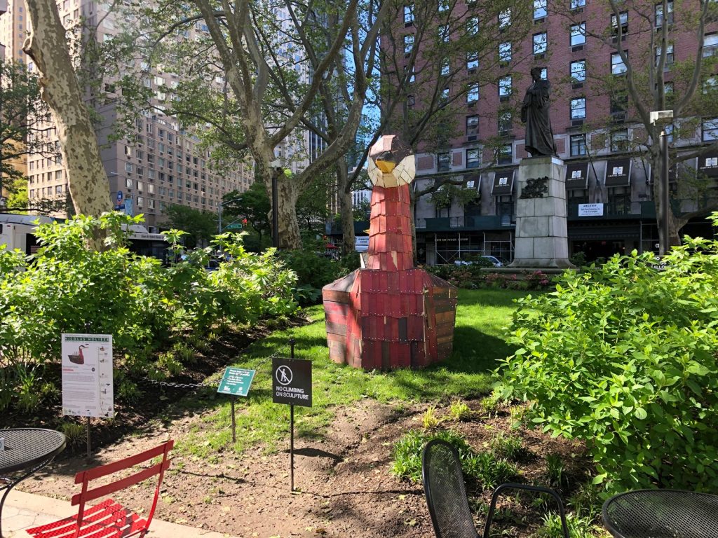a wooden sculpture of a red necked grebe sits on the grass area of Dante Park