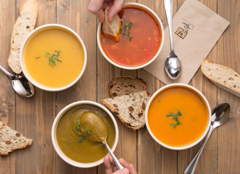 three soups on a wooden cutting board at Le Pain Quotidien with slices of bread and a hand dipping bread into one bowl